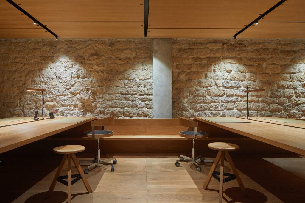 Büroeinrichtung mit Tischen, Stühlen und Hockern vor einem alten Steingemäuer.