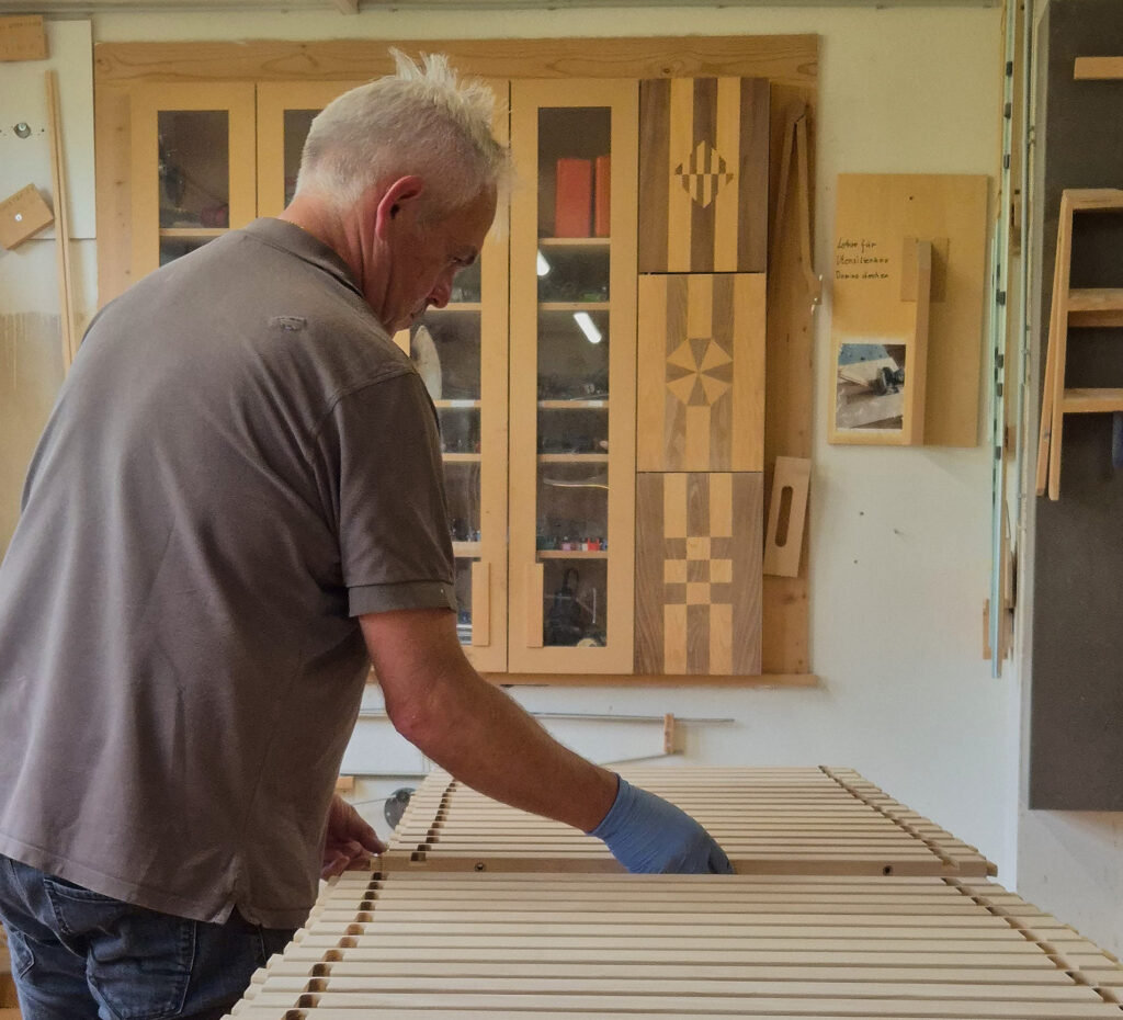 Handwerker in Werkstatt arbeitet an Tischbeinen, im Hintergrund Holzpaneele mit geometrischen Mustern an der Wand.